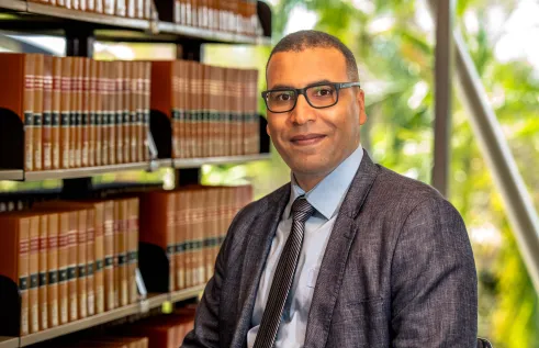 A man wearing glasses, a grey suit jacket, light blue shirt, and striped tie sits in a library with shelves of books behind him. He is smiling slightly and looking at the camera, with greenery visible through the window in the background.